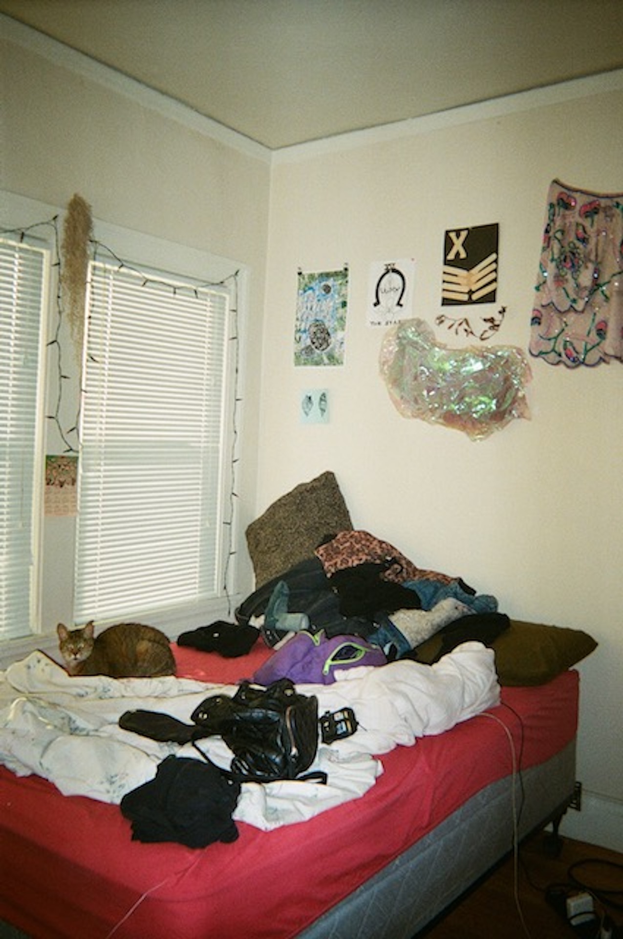 A bedroom with a messy bed and red bed sheets, light shining in through the window and various wall decorations